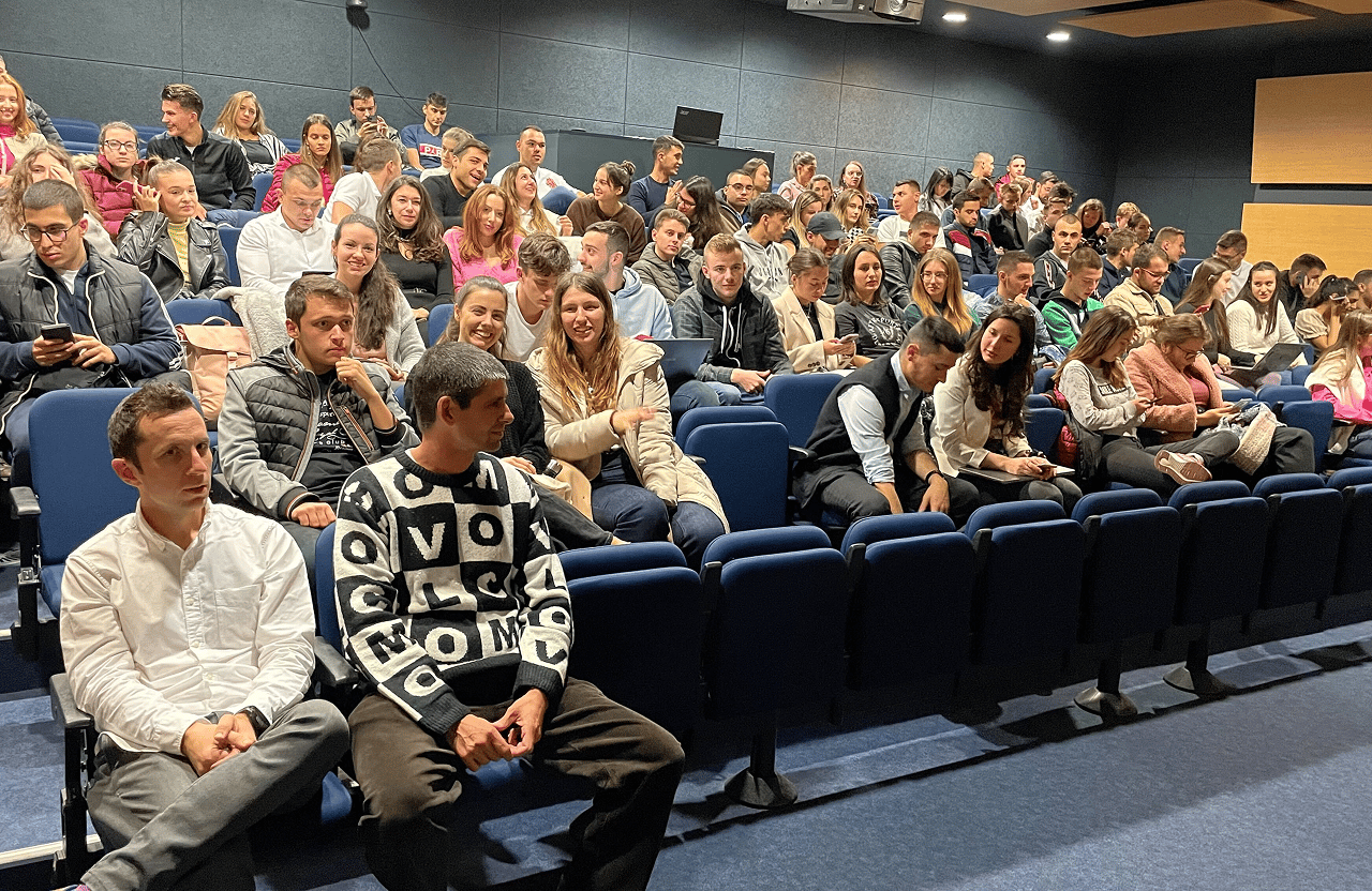 Large group of young adults seated in an auditorium, some using laptops and phones.