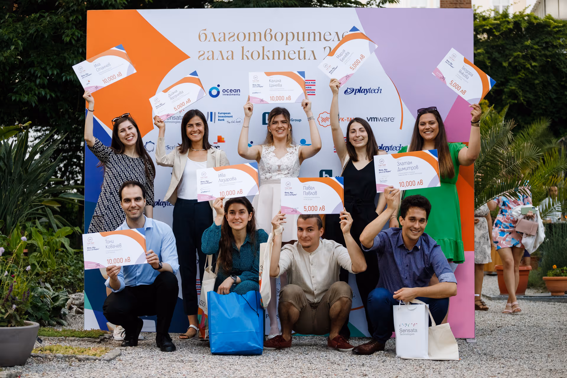 Group of nine smiling people holding oversized award checks at an outdoor gala event. Tuk-Tam scholars from 2022
